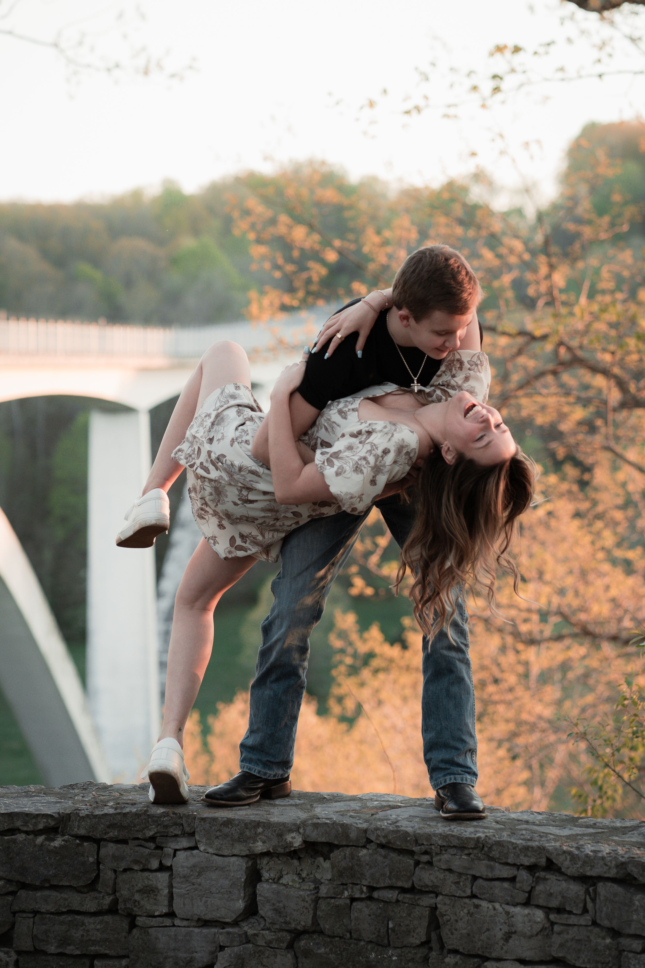 A romantic couple share a passionate dip kiss against a rustic stone wall at sunset with autumn trees in the background.