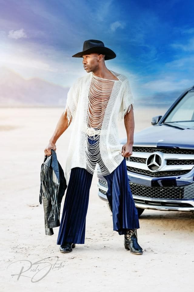 Fashion photo of model in white mesh top and blue pants posing with Mercedes Benz in desert setting.