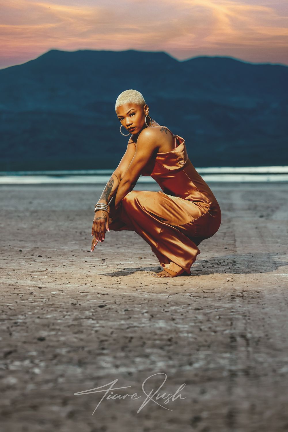 Figure in a flowing rust-colored dress crouches dramatically against a mountainous sunset backdrop.