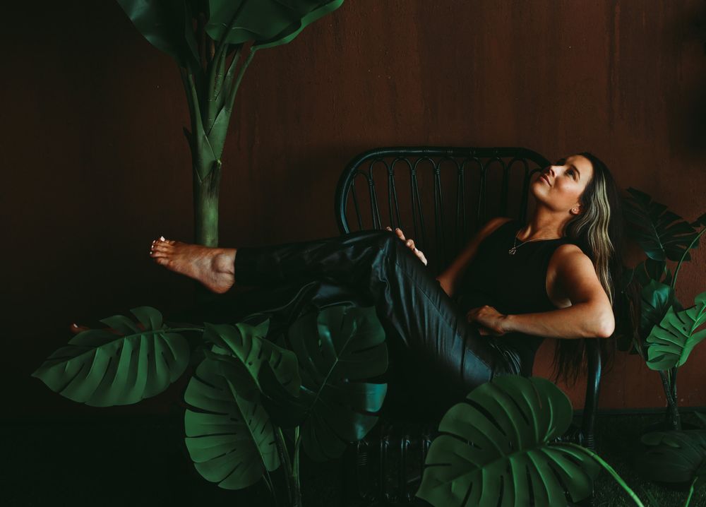 Artistic portrait with green tropical leaves showing a figure lounging on dark furniture in moody lighting.