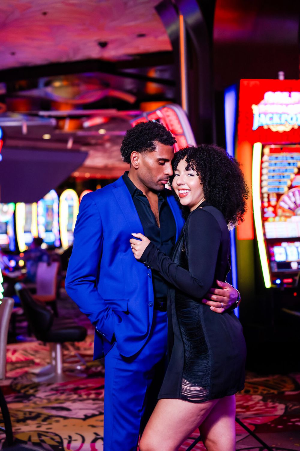 Stylish couple embracing at a casino with neon lights, wearing blue suit and black dress.