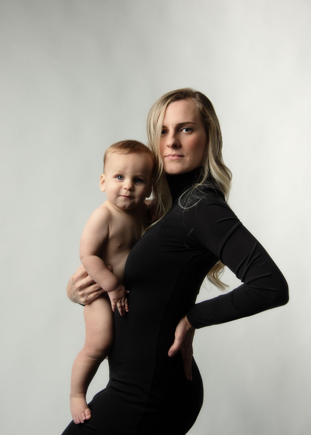 Mother in black turtleneck poses with nude baby against minimal backdrop.
