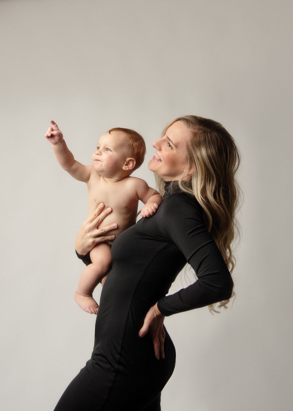 Mother in black holds smiling baby who points upward against neutral background.