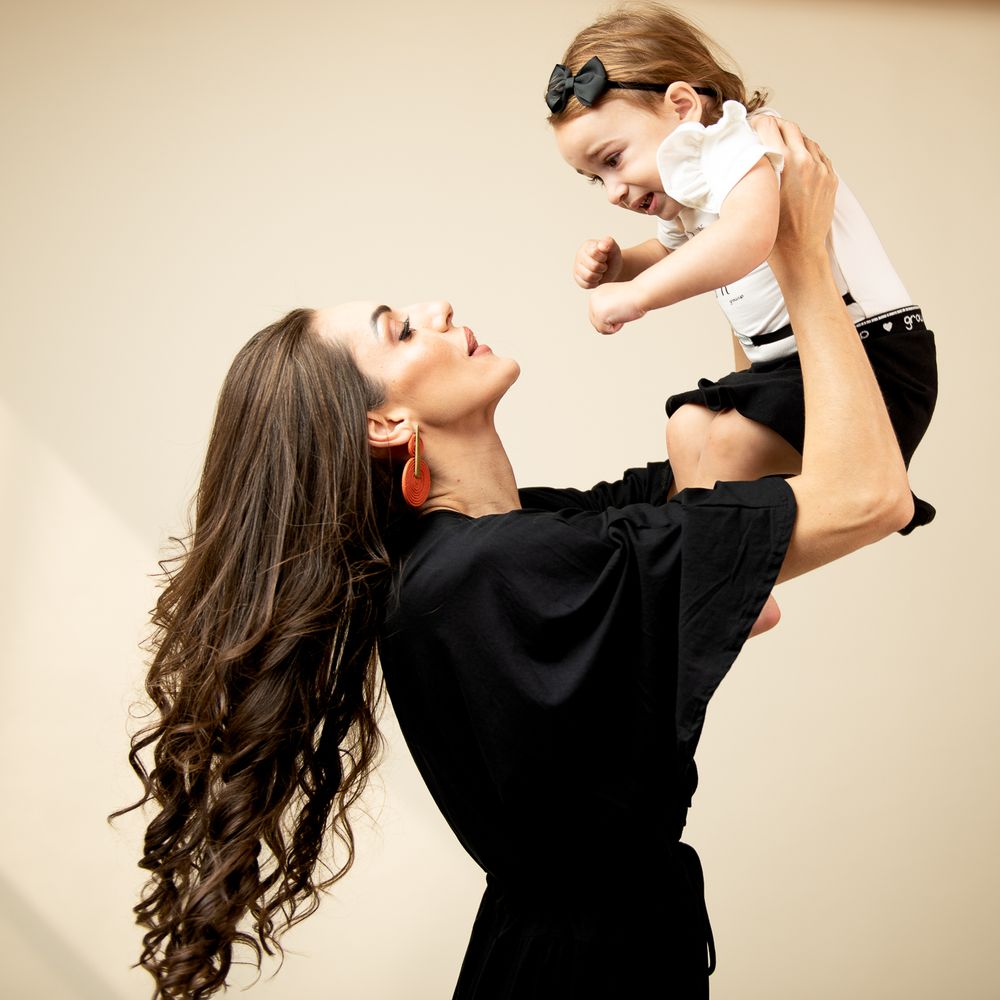 A parent in black dress holds and lifts smiling baby wearing white outfit in studio setting.