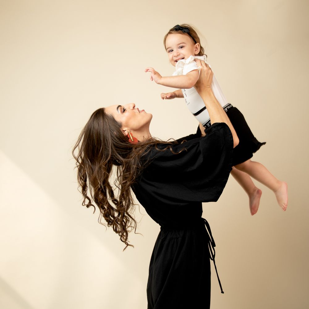A parent in black dress lifts and plays with baby in white outfit against beige background.