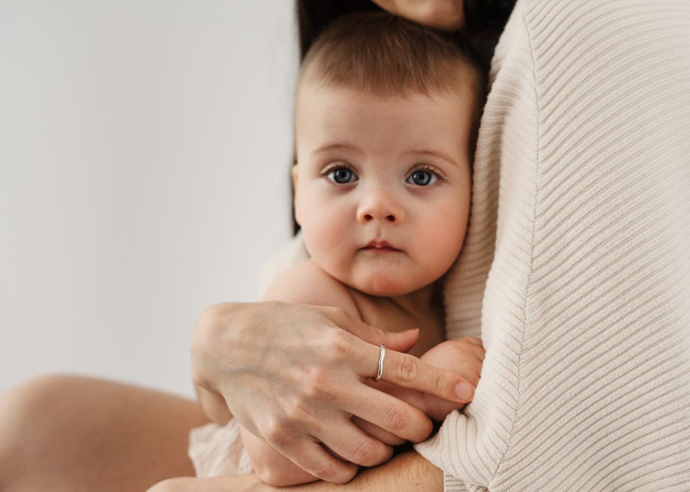 Close-up of a baby in beige clothing looking directly at camera while being held.