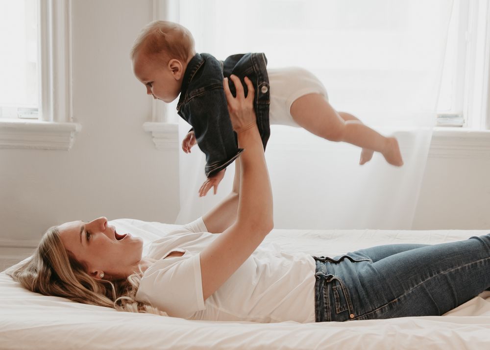A joyful parent lying on bed lifts and plays with a baby wearing dark clothing against a bright white background.