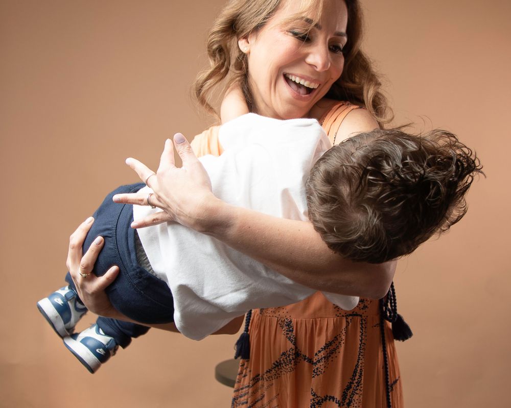 Mother in patterned dress playfully holds laughing child upside down.