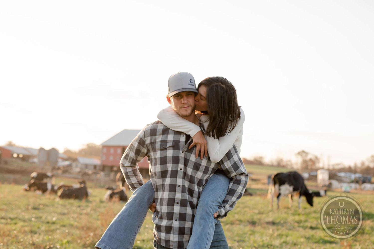 A playful piggyback ride moment captured on a scenic farm with grazing cattle nearby.