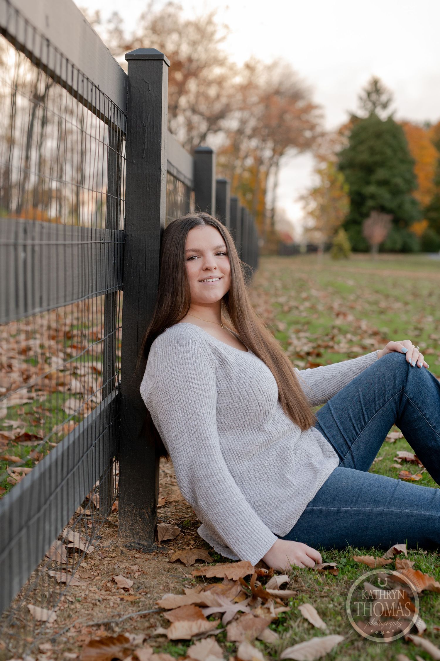 A figure relaxes against a metal fence on a ground covered with fallen autumn leaves.
