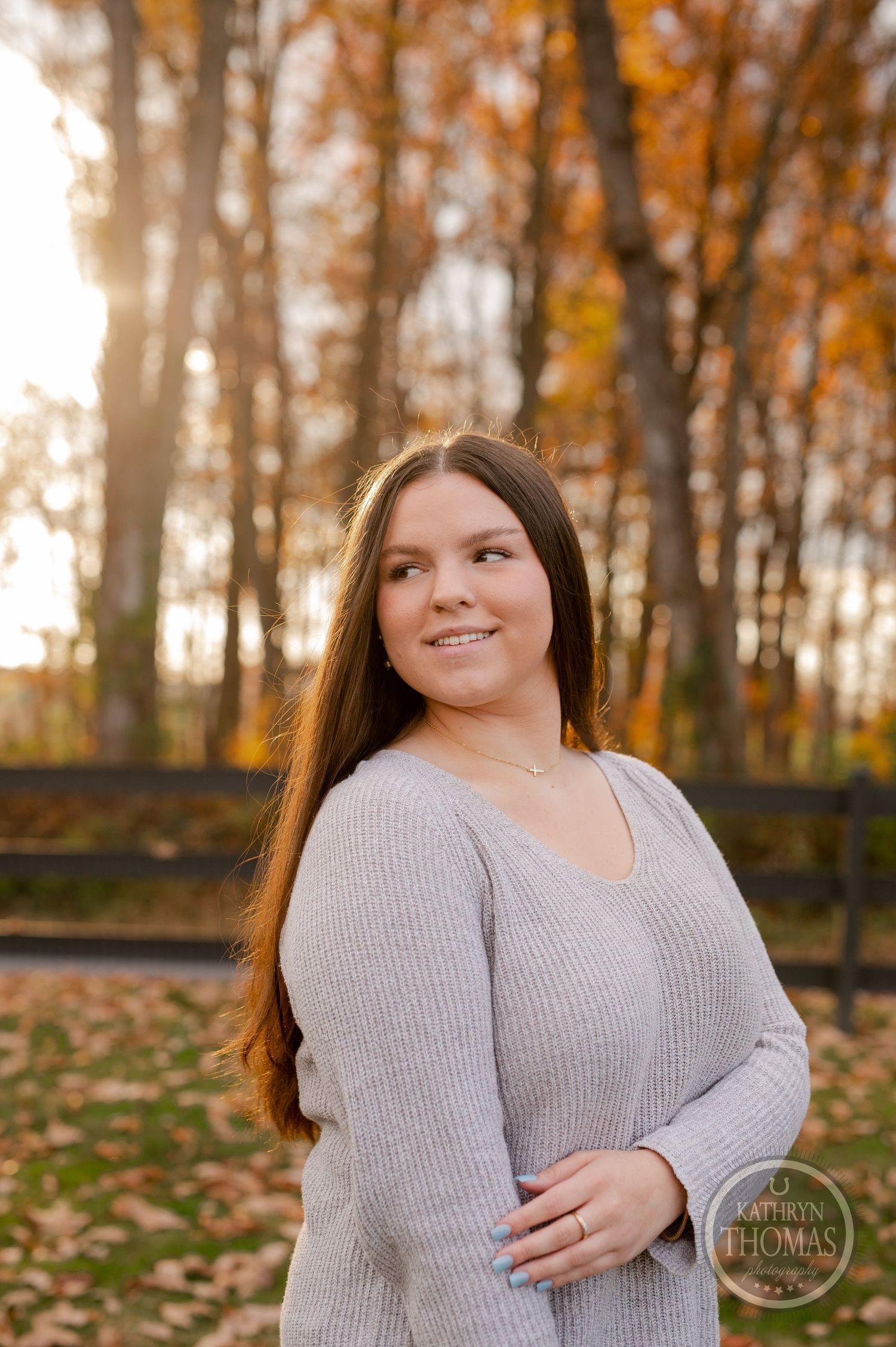Fall portrait in a warm sweater with golden autumn trees and wooden fence in background.