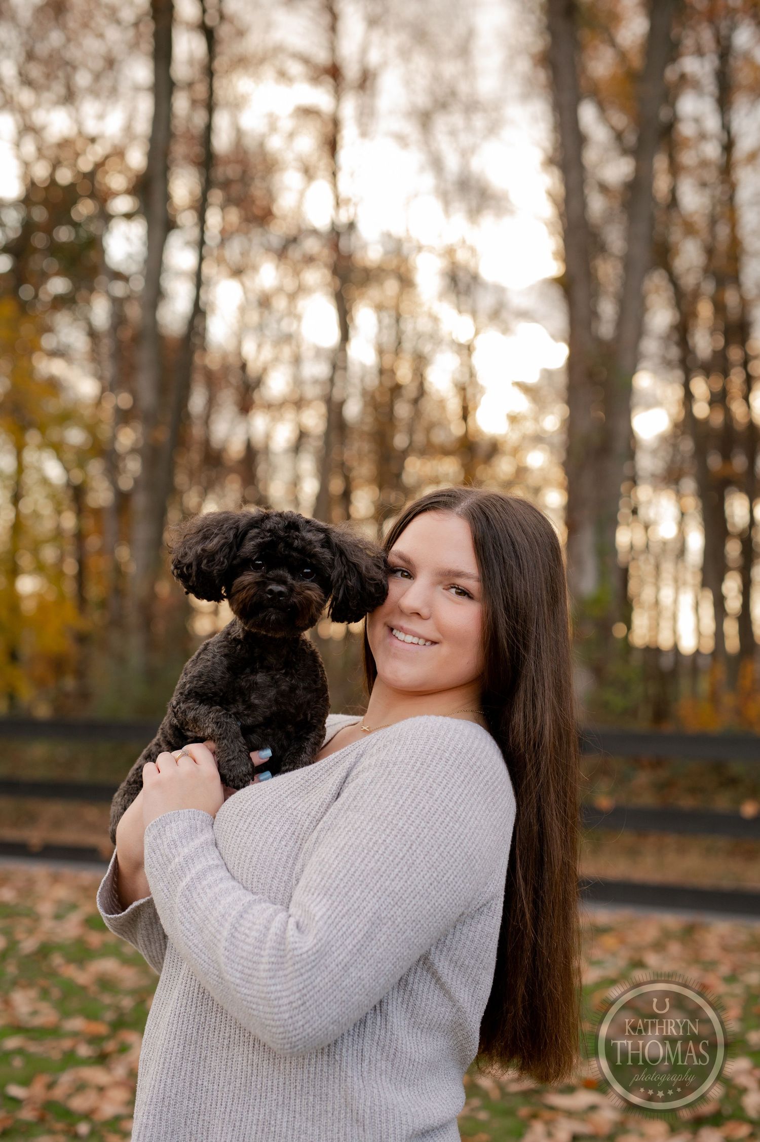 Person in grey sweater holds a small black dog against fall foliage and wooden fence.