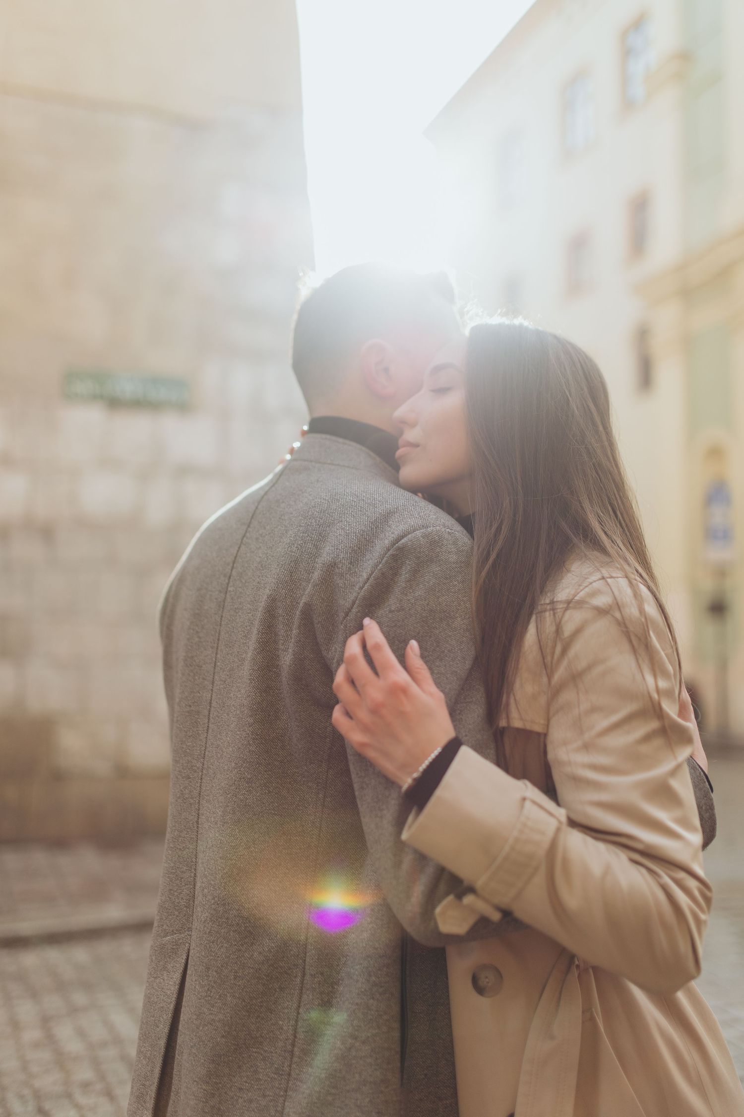 Intimate couple embracing in sunlit cobblestone street wearing matching trench coats.