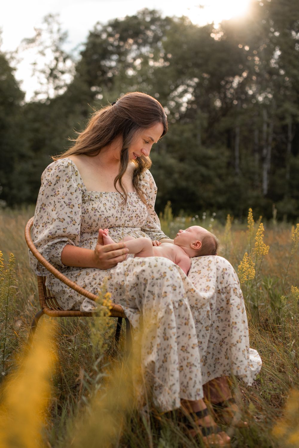 Mother and newborn baby in floral dress amid golden wildflowers at sunset.