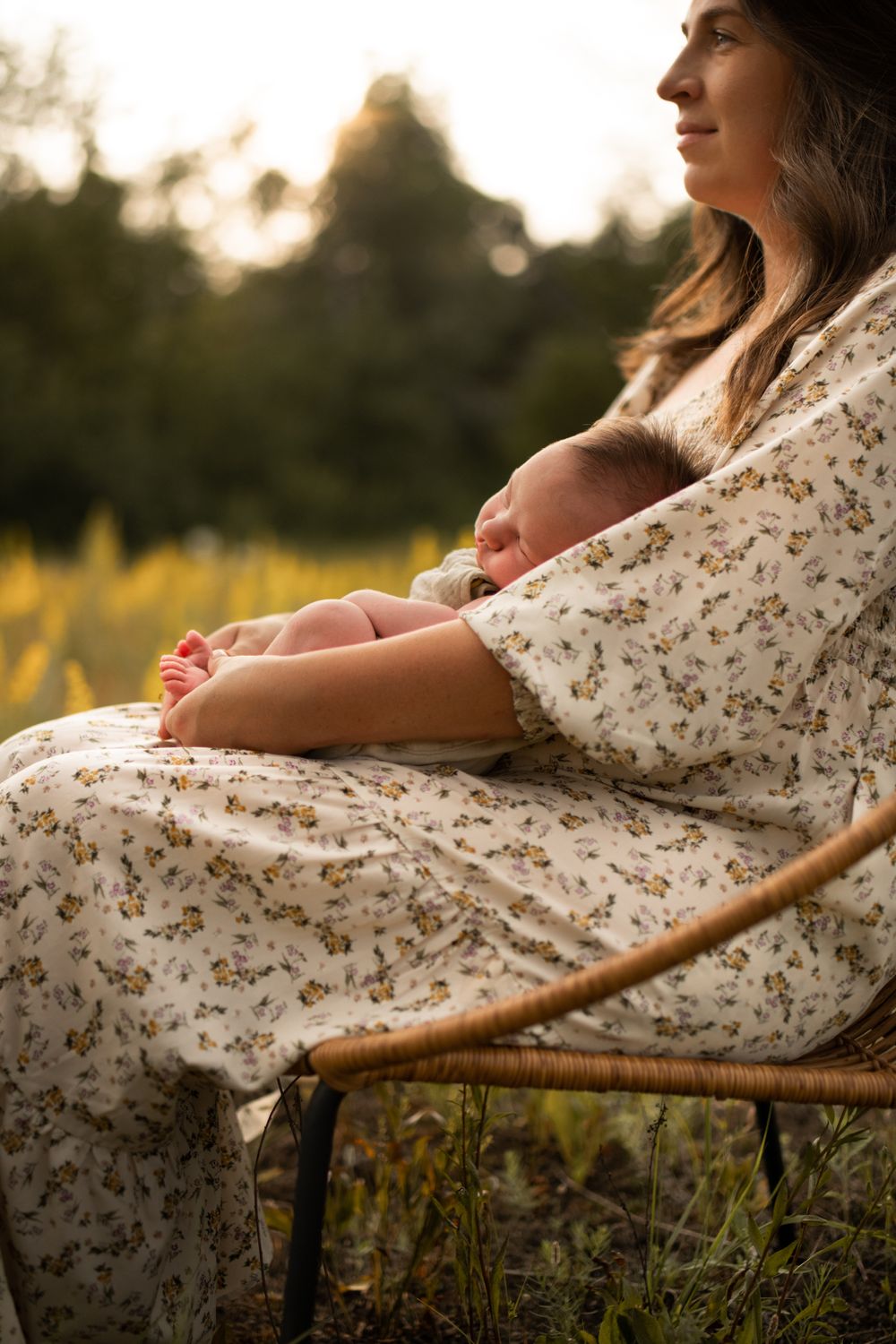 Mother wearing a floral dress resting with an infant outdoors during golden hour.