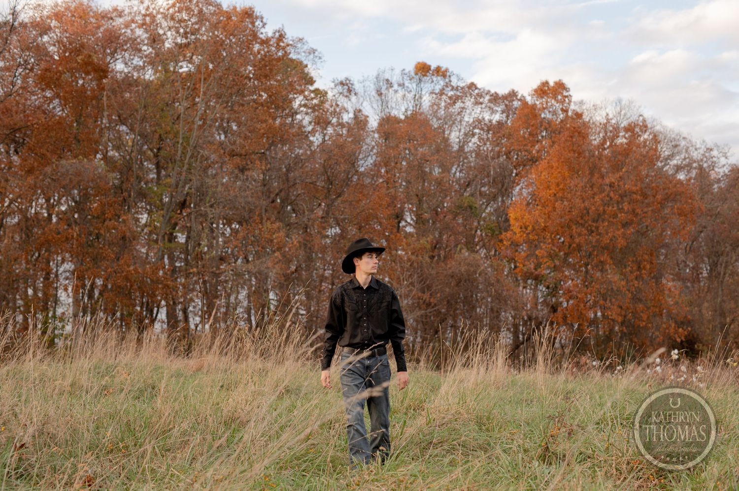 A figure in black attire walks through an autumn field with orange trees in the background.