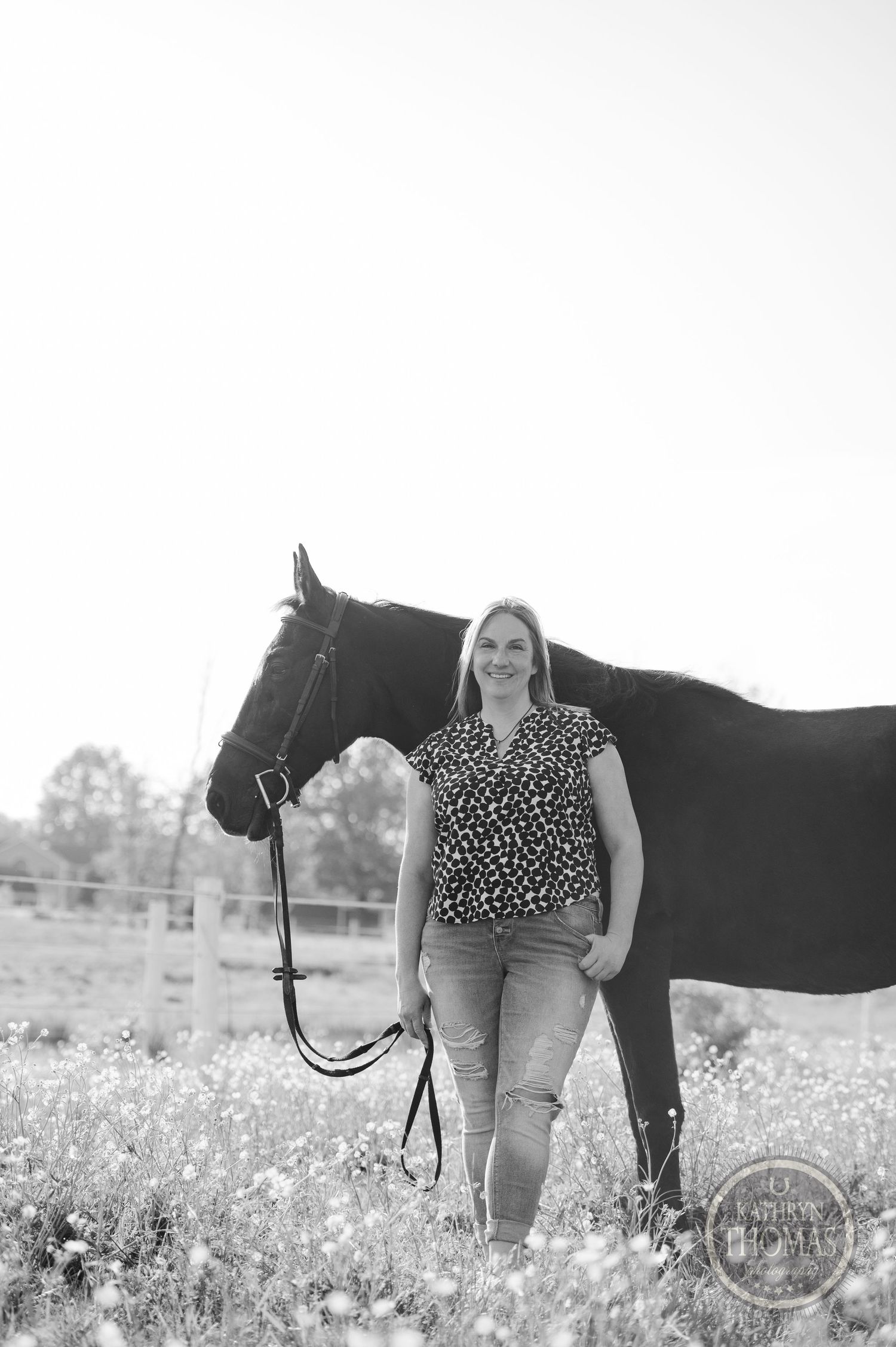 Black and white portrait of person standing with black horse in wildflower field.