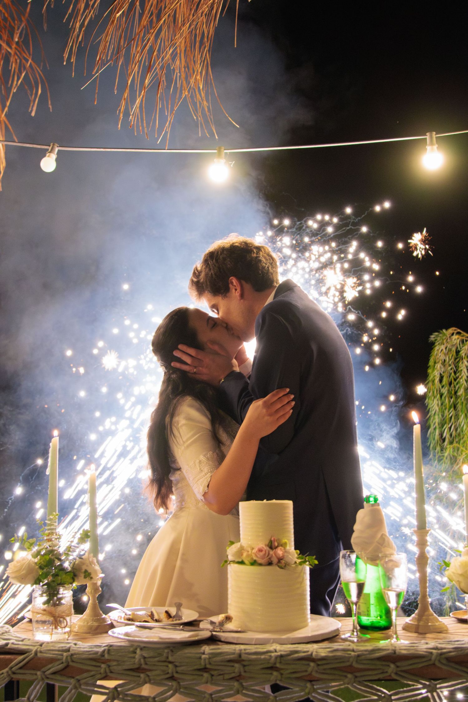 A magical wedding kiss with sparklers and string lights illuminating the reception dessert table.