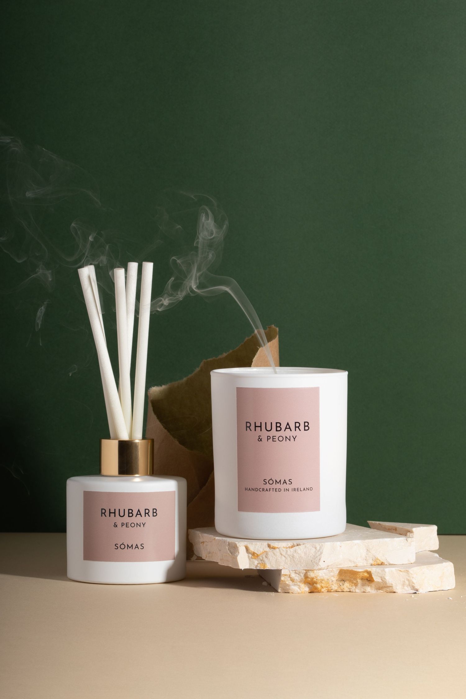 Rhubarb & Peony reed diffuser and candle emitting smoke against dark green background. Placed on tasteful broken stone.