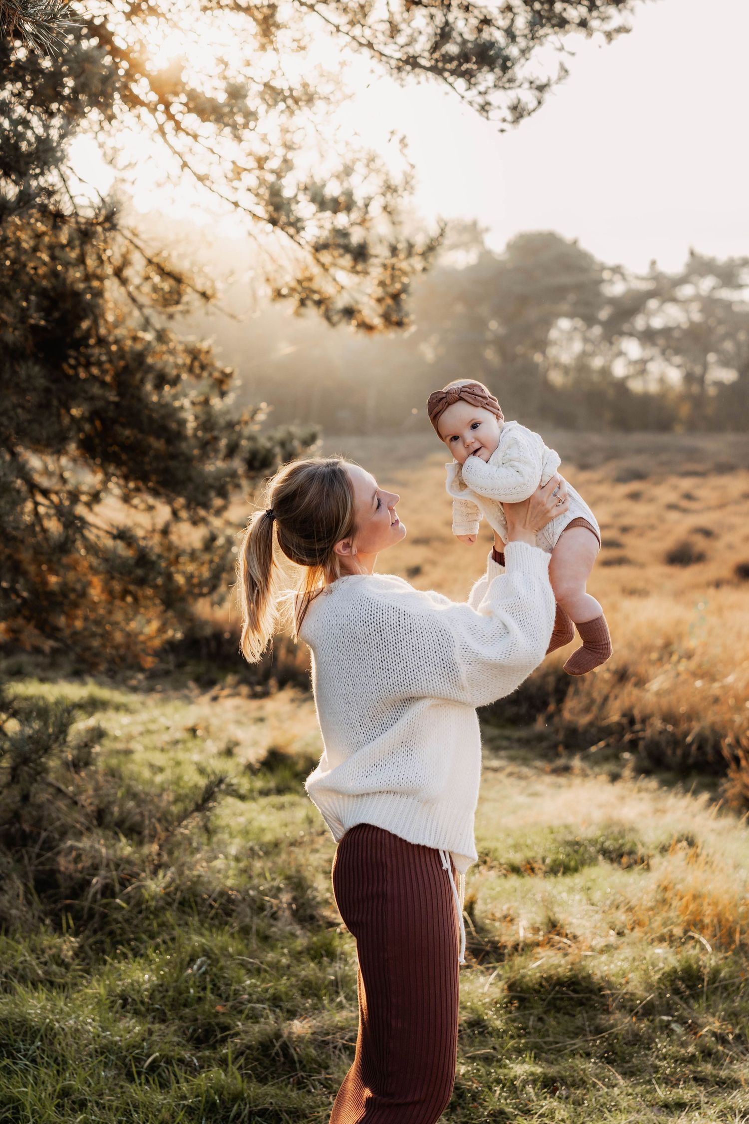 Mother lifting baby in white sweater during golden hour in nature.