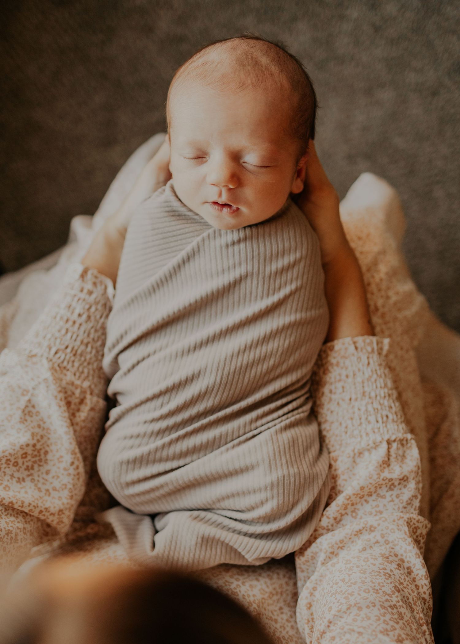 Peaceful sleeping newborn baby swaddled in gray fabric resting on soft beige blanket in warm lighting.