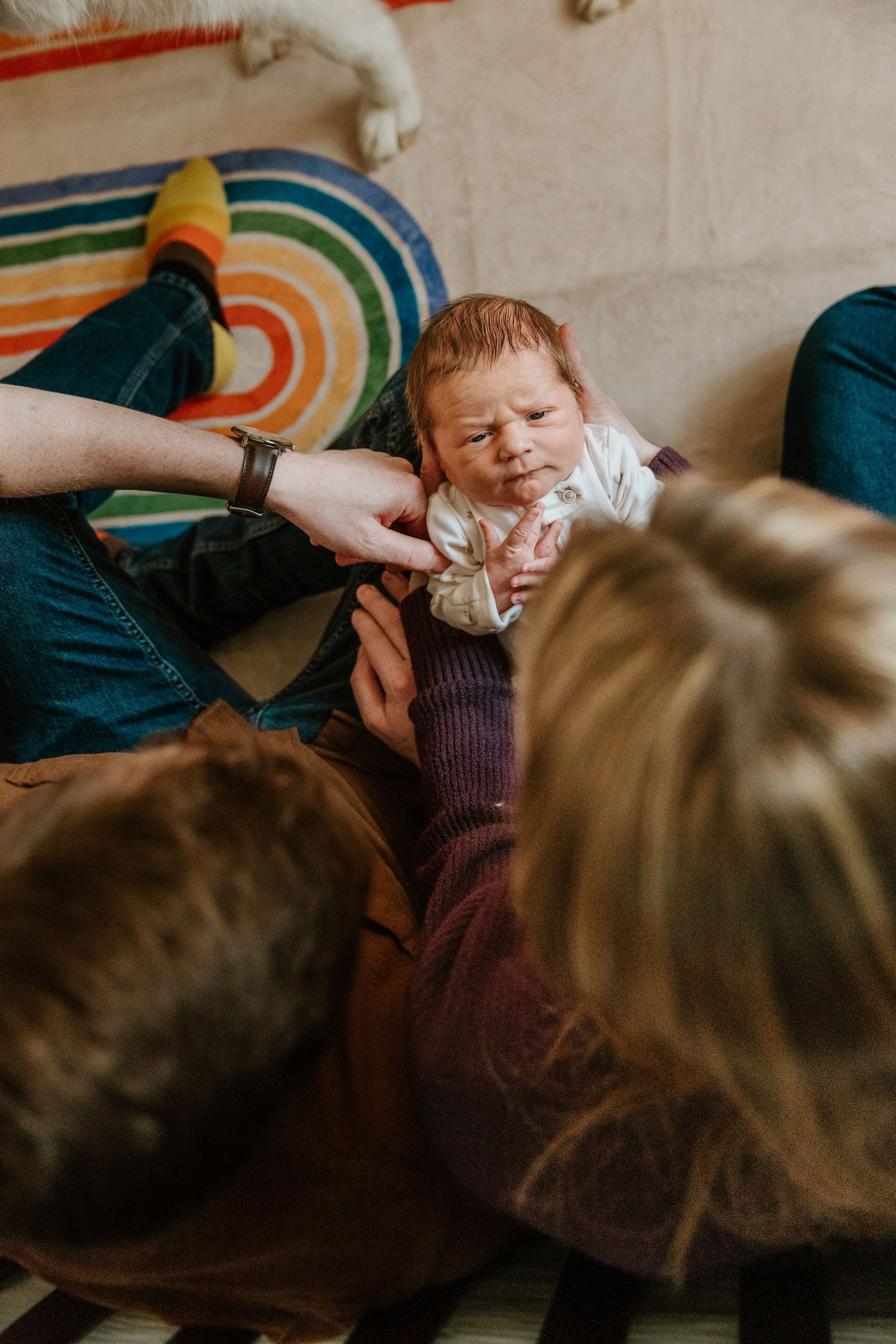Two parents lovingly hold their newborn baby with red hair on a colorful rainbow rug at home.
