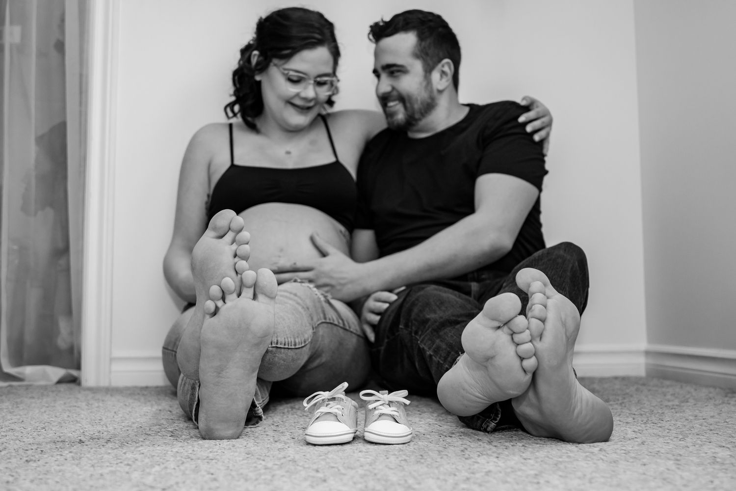 A black and white photograph showing an expectant couple sitting on carpet with tiny baby shoes between their bare feet.
