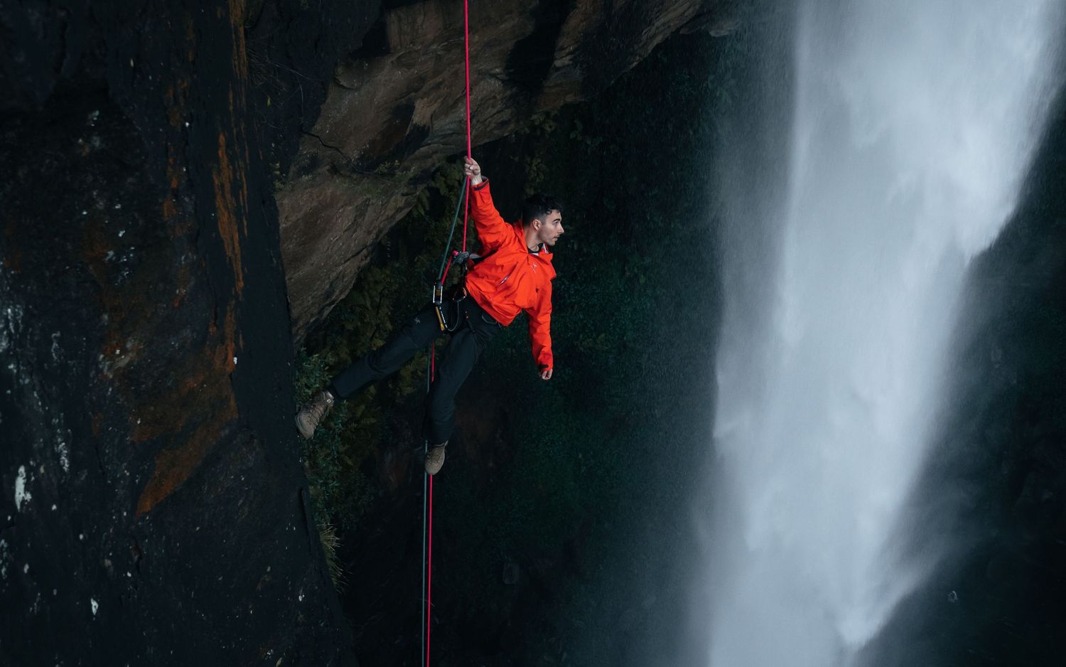 Rock climber in red jacket rappels down dramatic waterfall cliffs on a single rope.