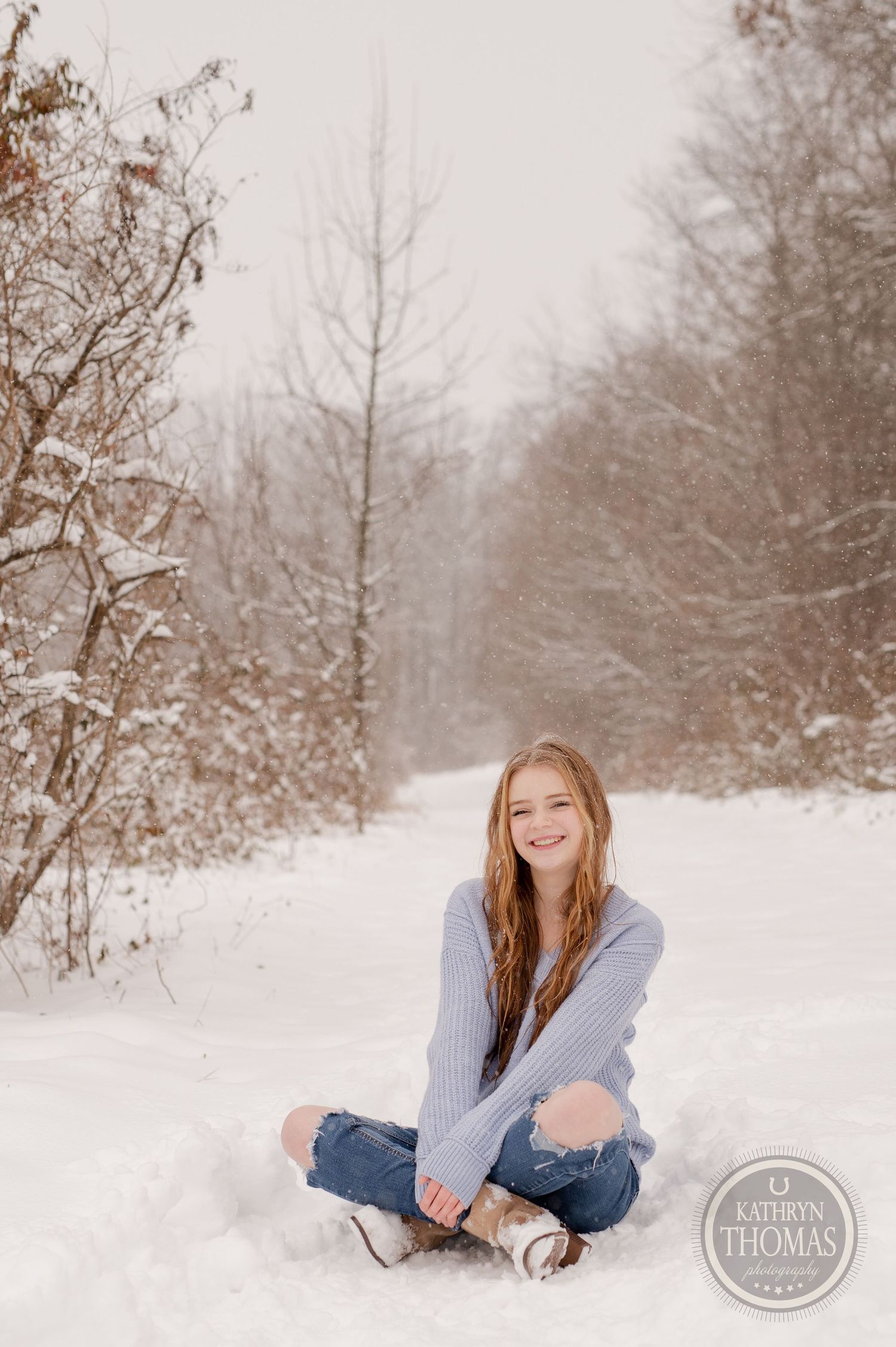 A person sitting in snow wearing casual winter attire smiles on a snowy forest trail.