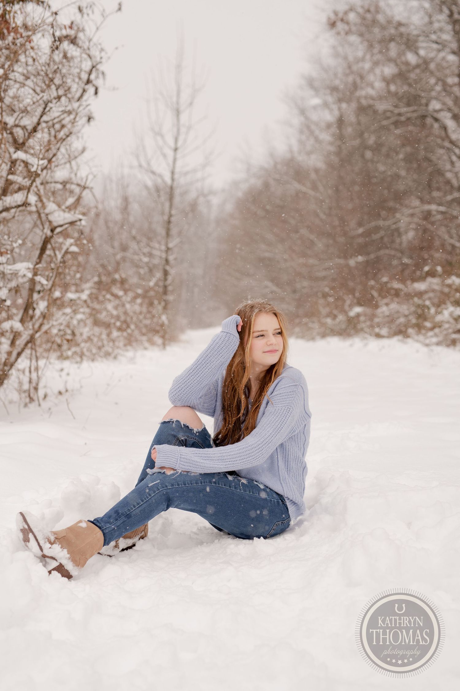A person reclines in fresh snow wearing a gray sweater and jeans on a winter trail.