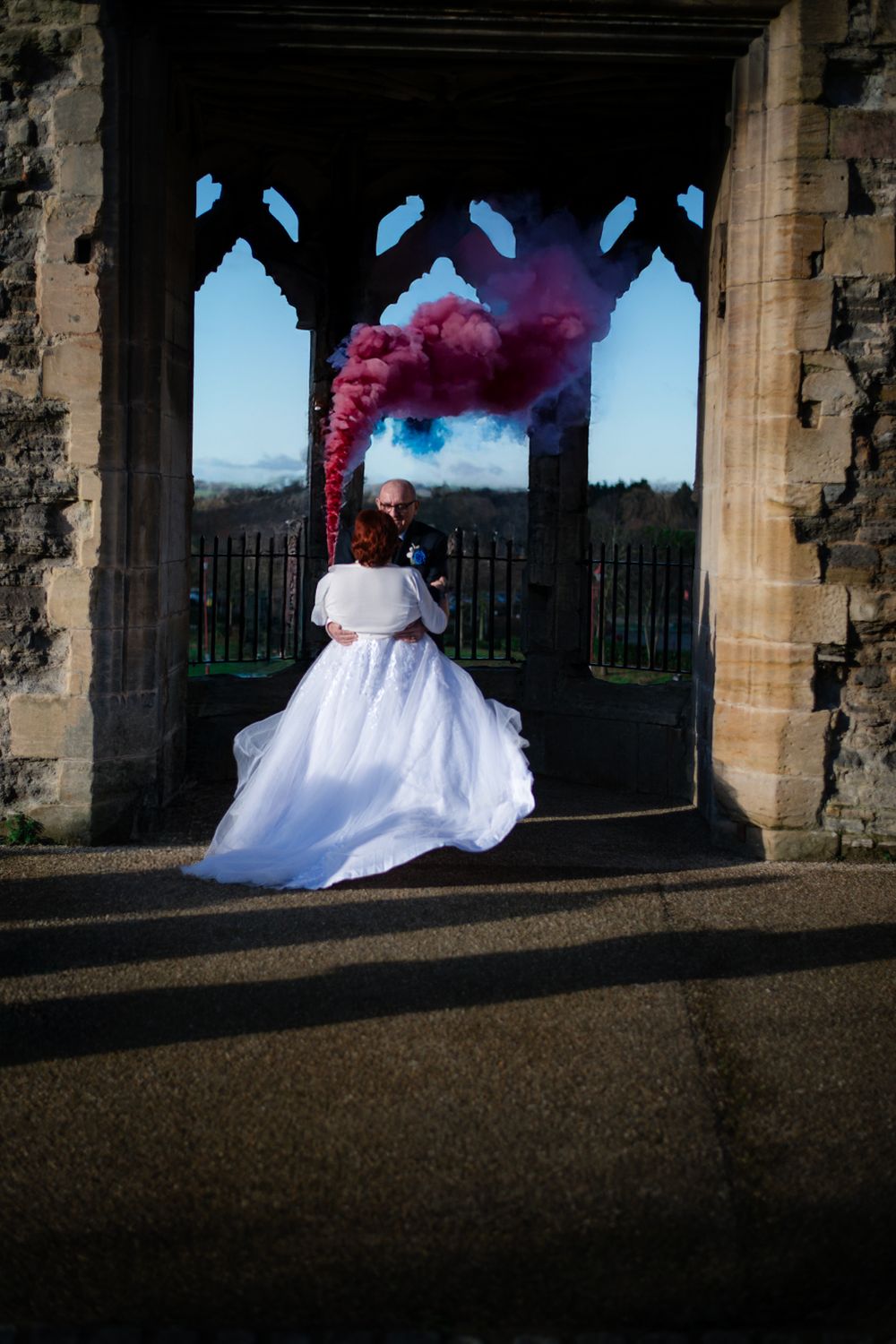 Wedding photo with pink smoke bombs creating dramatic effect under gothic stone archway.