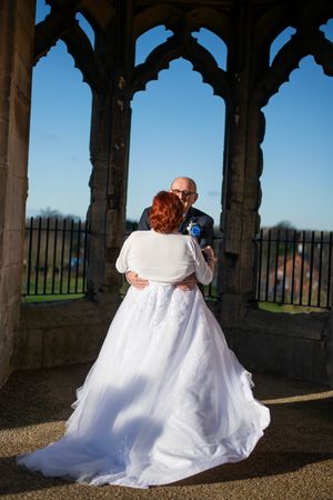 Couple in wedding attire poses under gothic stone arches with blue sky backdrop.
