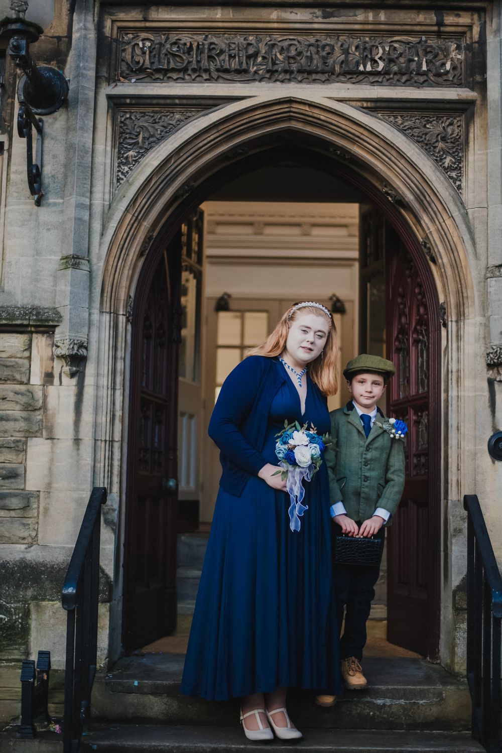 Wedding party members in navy blue dress and green suit standing in ornate stone doorway.