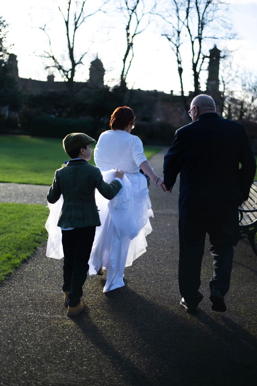 Wedding party walks along garden path with historic building silhouette in background.