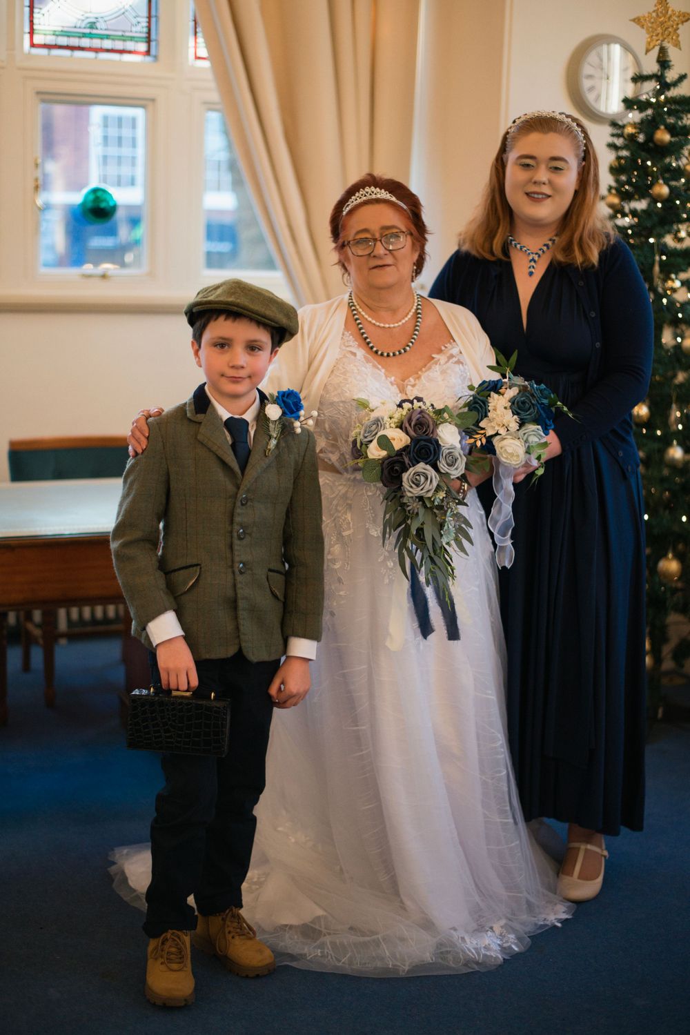 Wedding group photo with Christmas tree and elegant white dress against cream-colored walls.