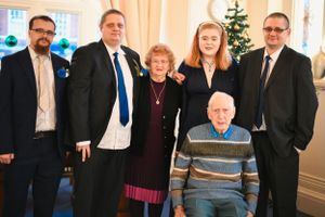 Family group poses in formal attire around antique desk with Christmas tree visible in background.
