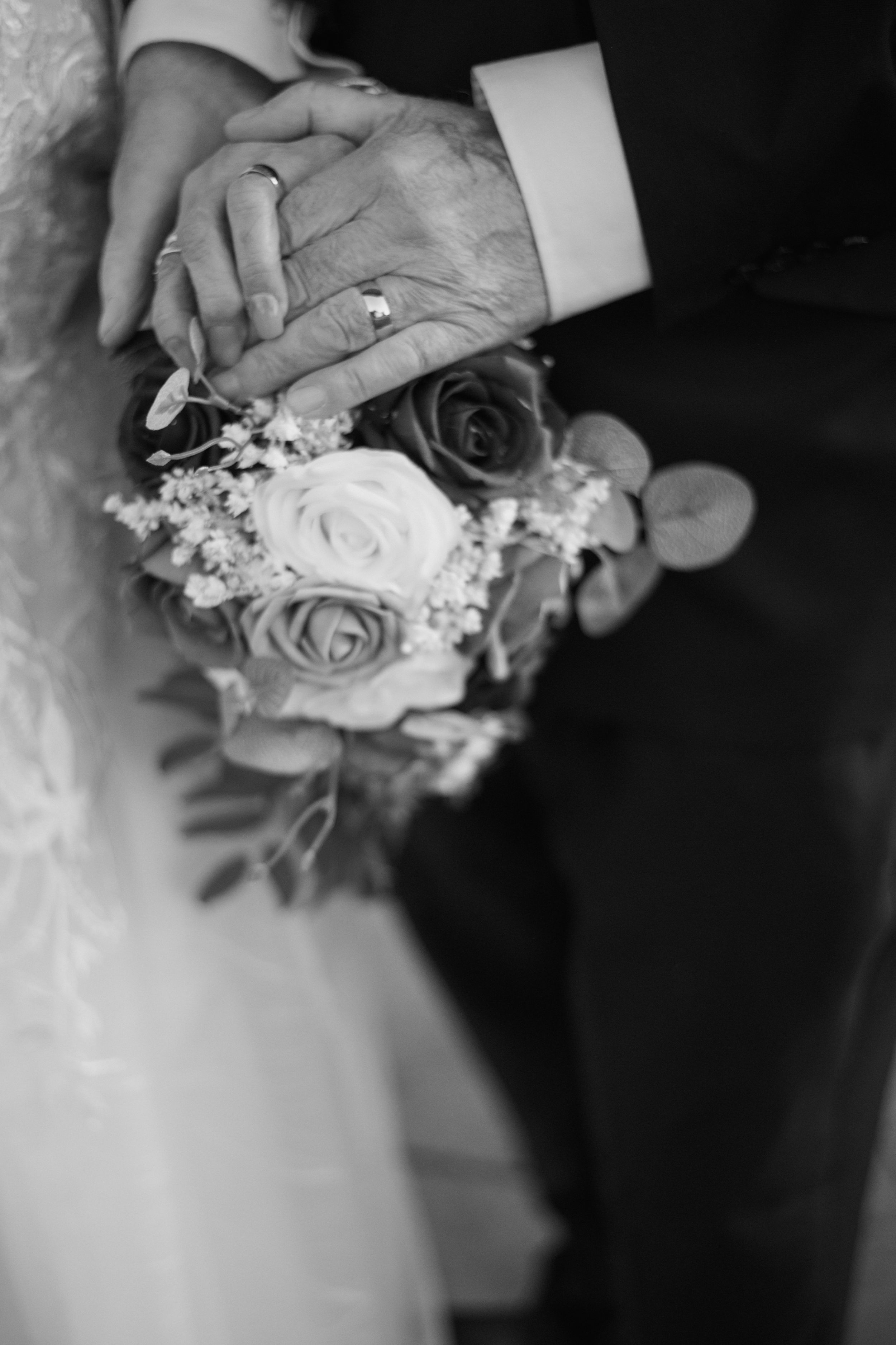 Close-up black and white photo of clasped hands holding a bouquet of roses with white lace details.