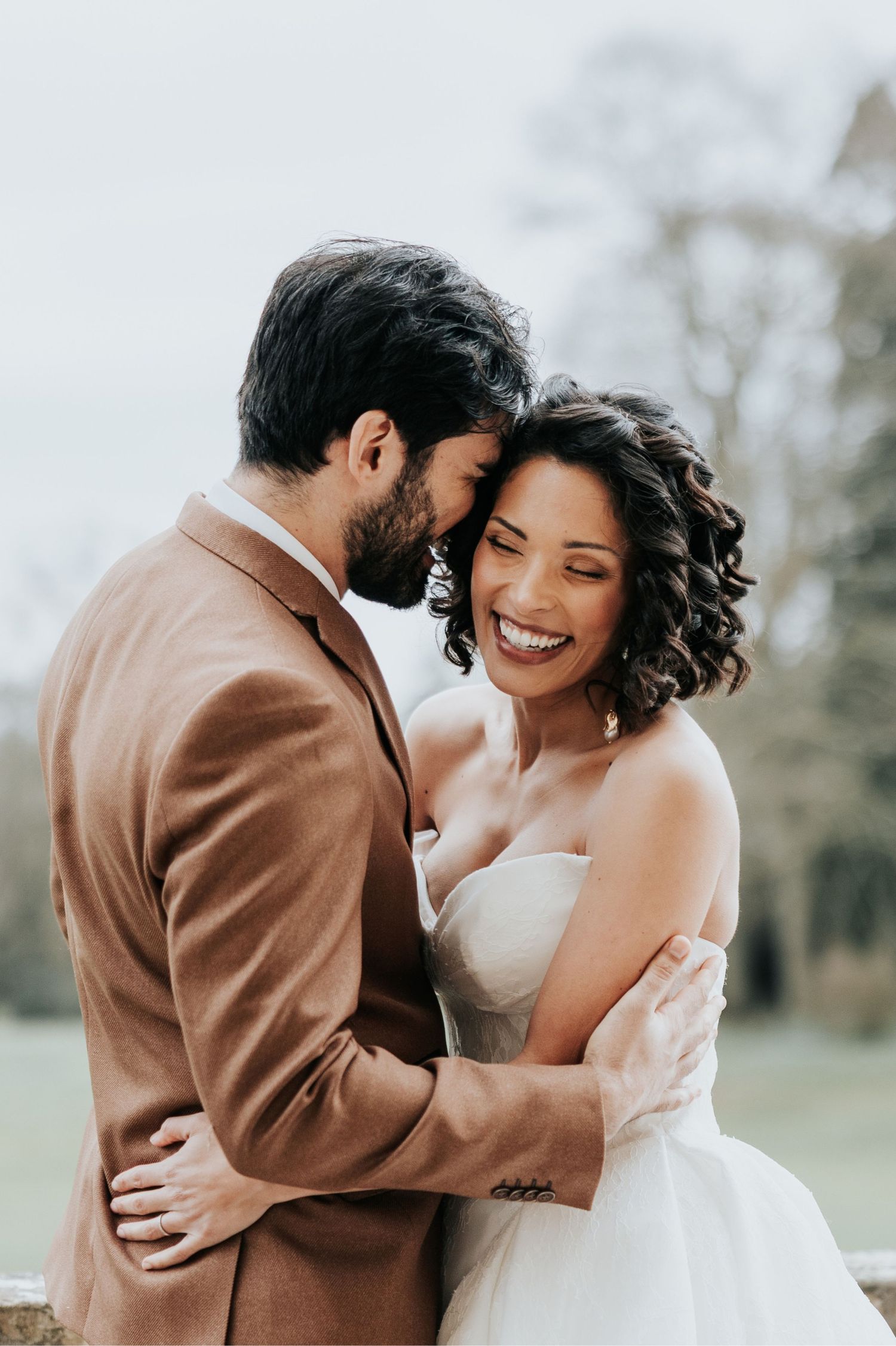 A joyful couple shares an intimate moment outdoors, with the bride in white and groom in brown embracing and smiling.