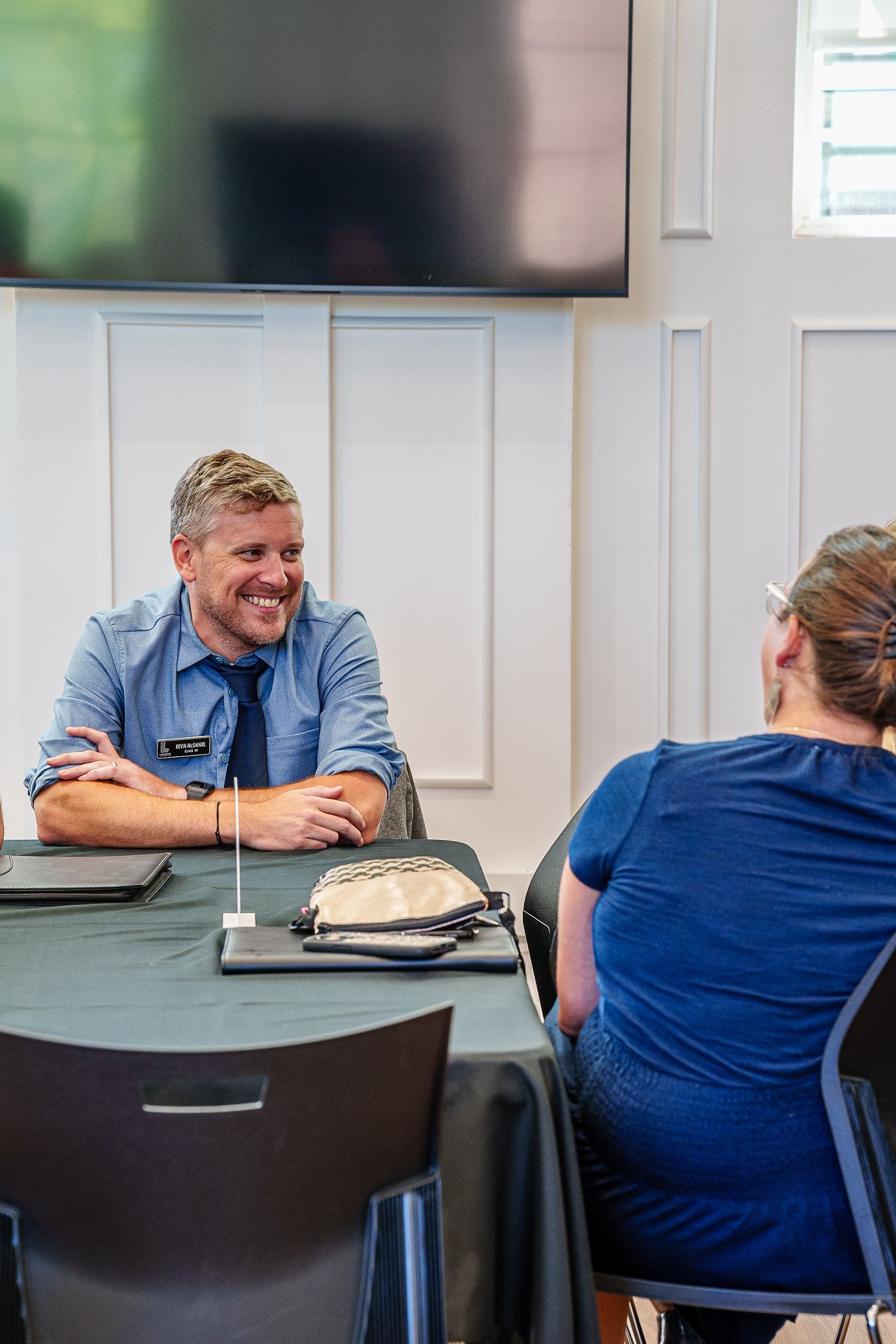 Two people in blue attire having an animated conversation at a meeting table with a flat screen TV mounted on the wall.