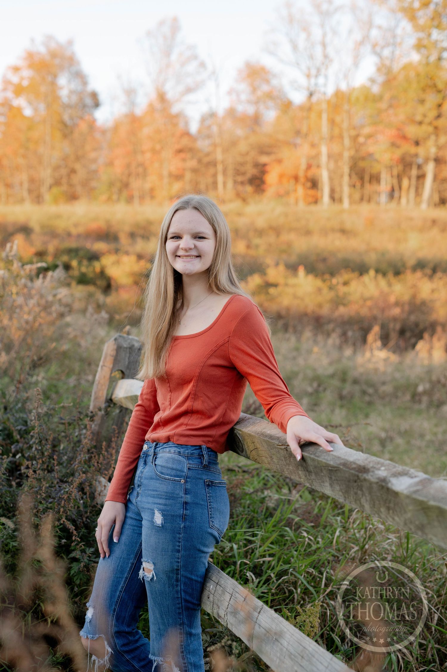 Person in coral sweater and ripped jeans poses by wooden fence in autumn field during golden hour sunset.