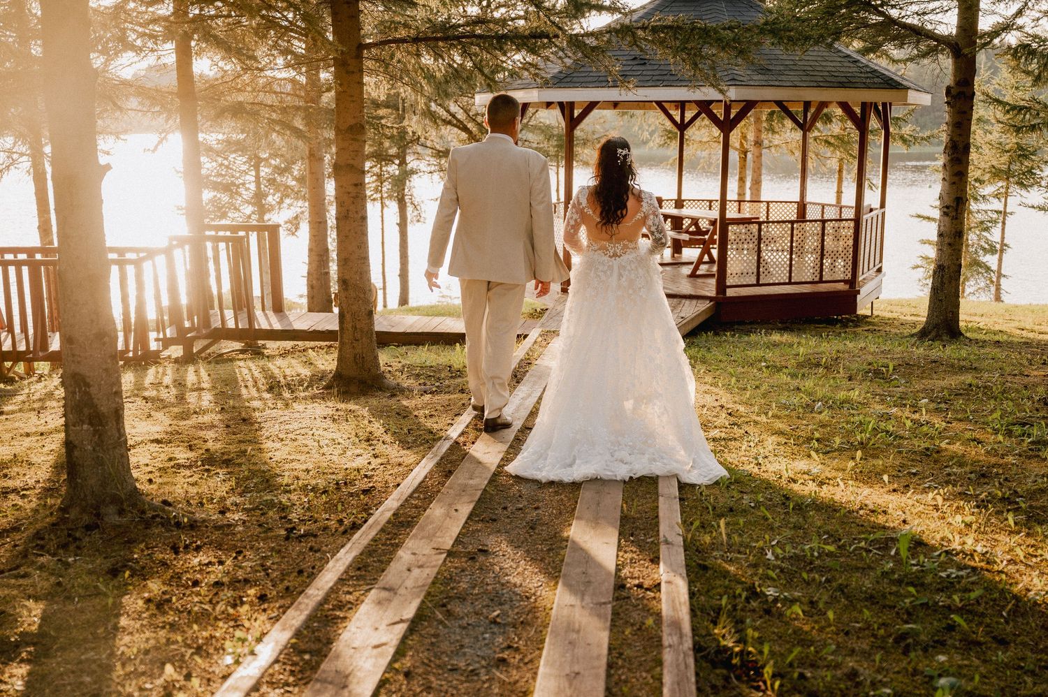 Newlyweds walk towards a lakeside gazebo during golden hour with long shadows on a wooded path.