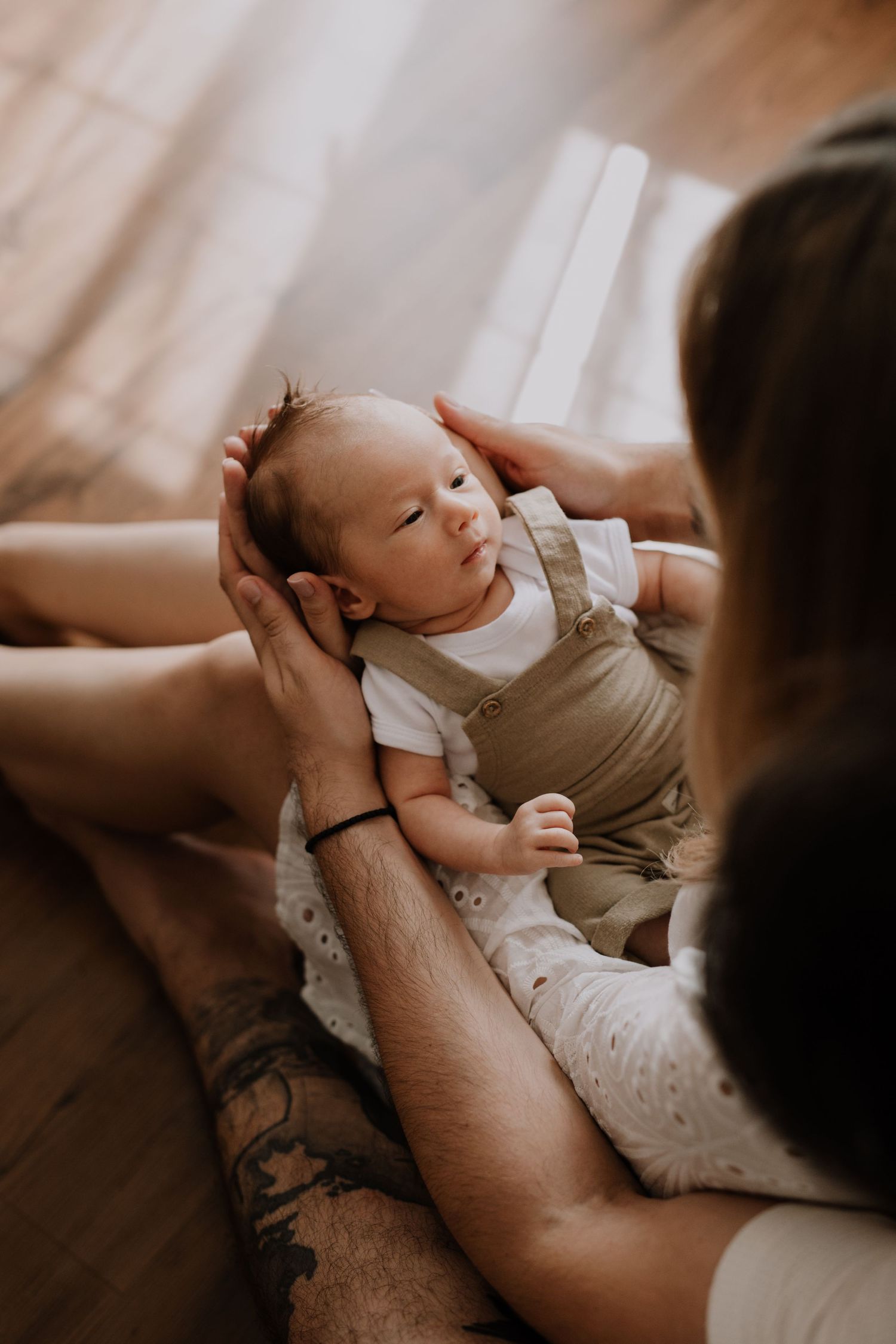 Baby resting peacefully while being held with tattooed arms in soft indoor lighting.