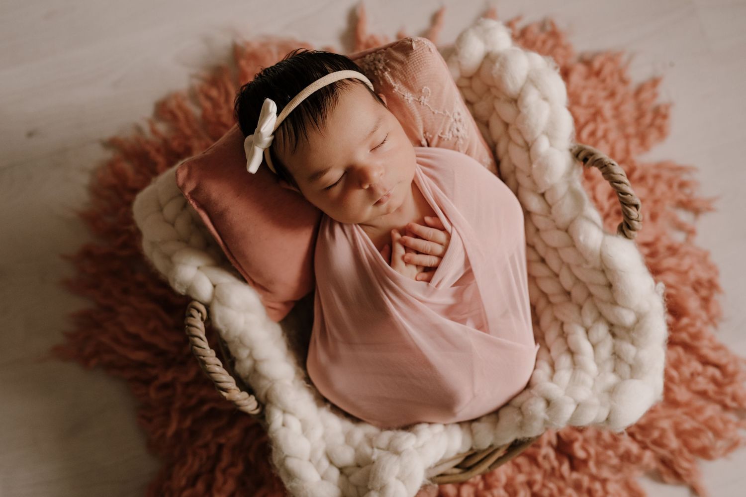 Sleeping newborn wrapped in pink swaddle on textured blankets with peach and cream tones.