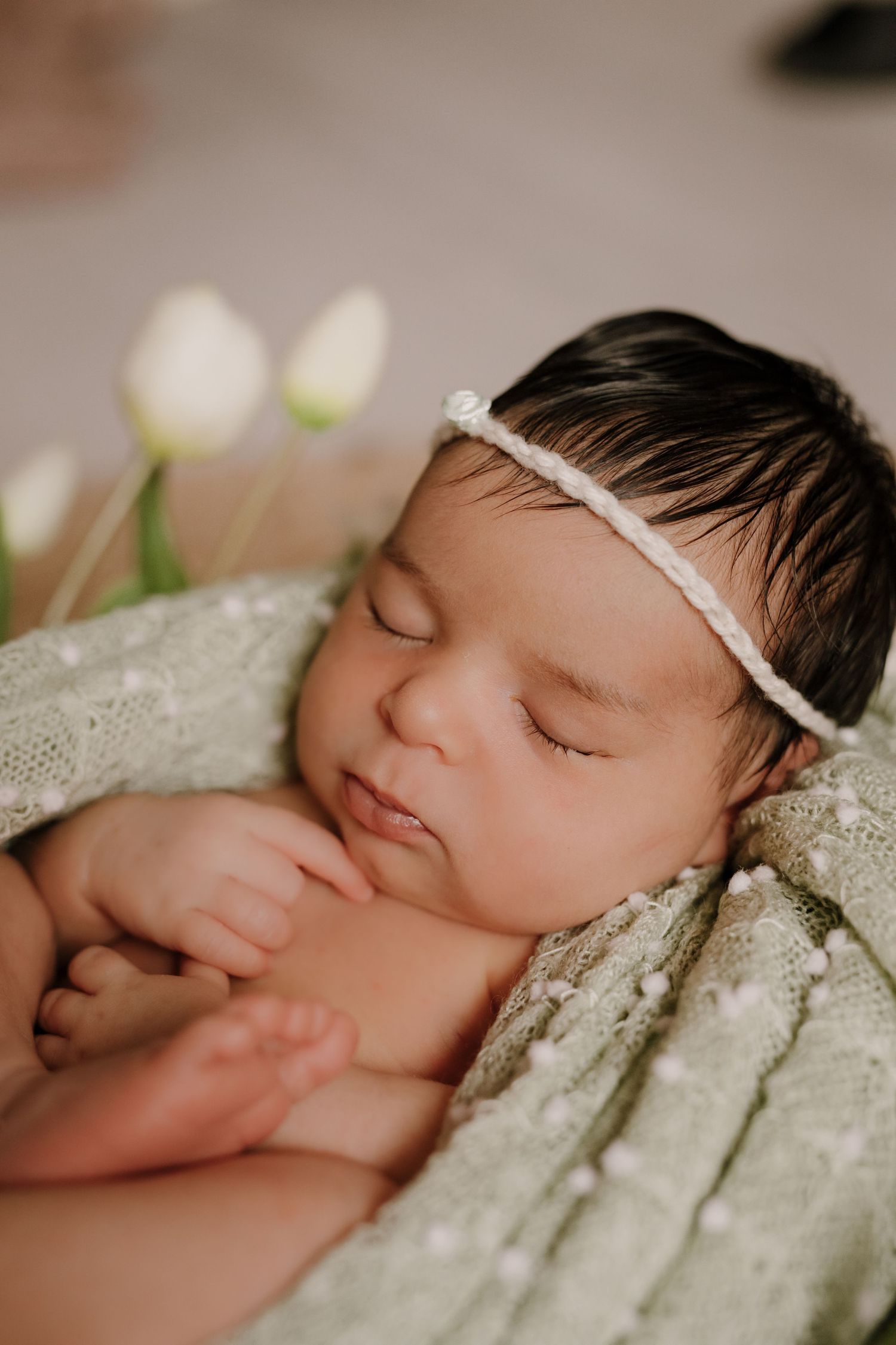 Sleeping newborn with delicate white headband on lace fabric near tulips.