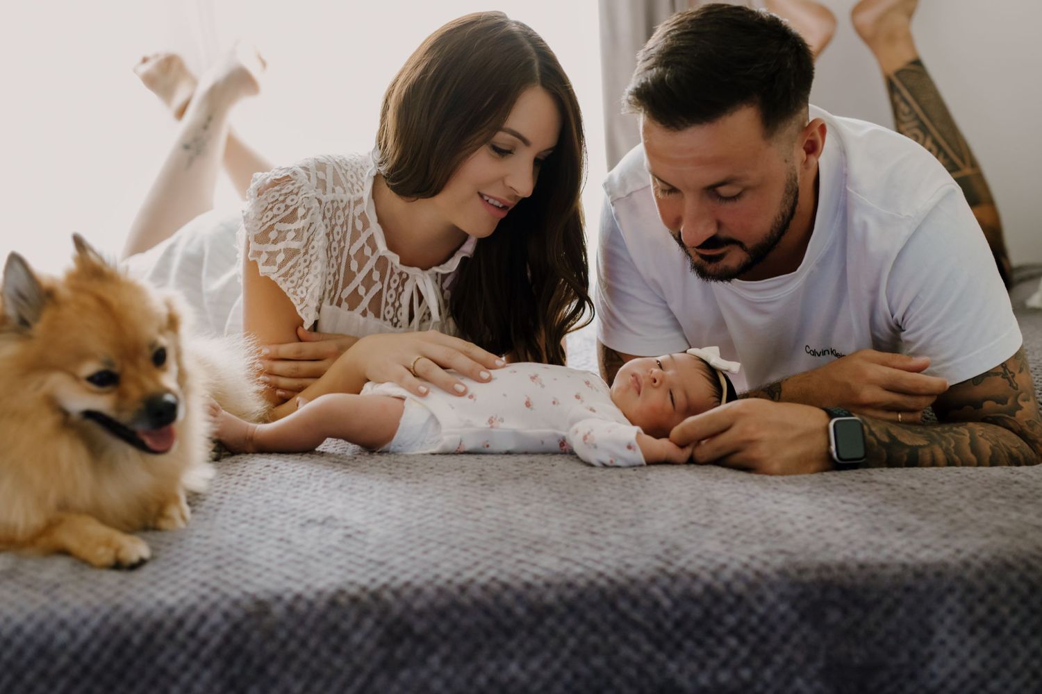 Parents admiring their newborn baby while a Pomeranian dog watches from the side.