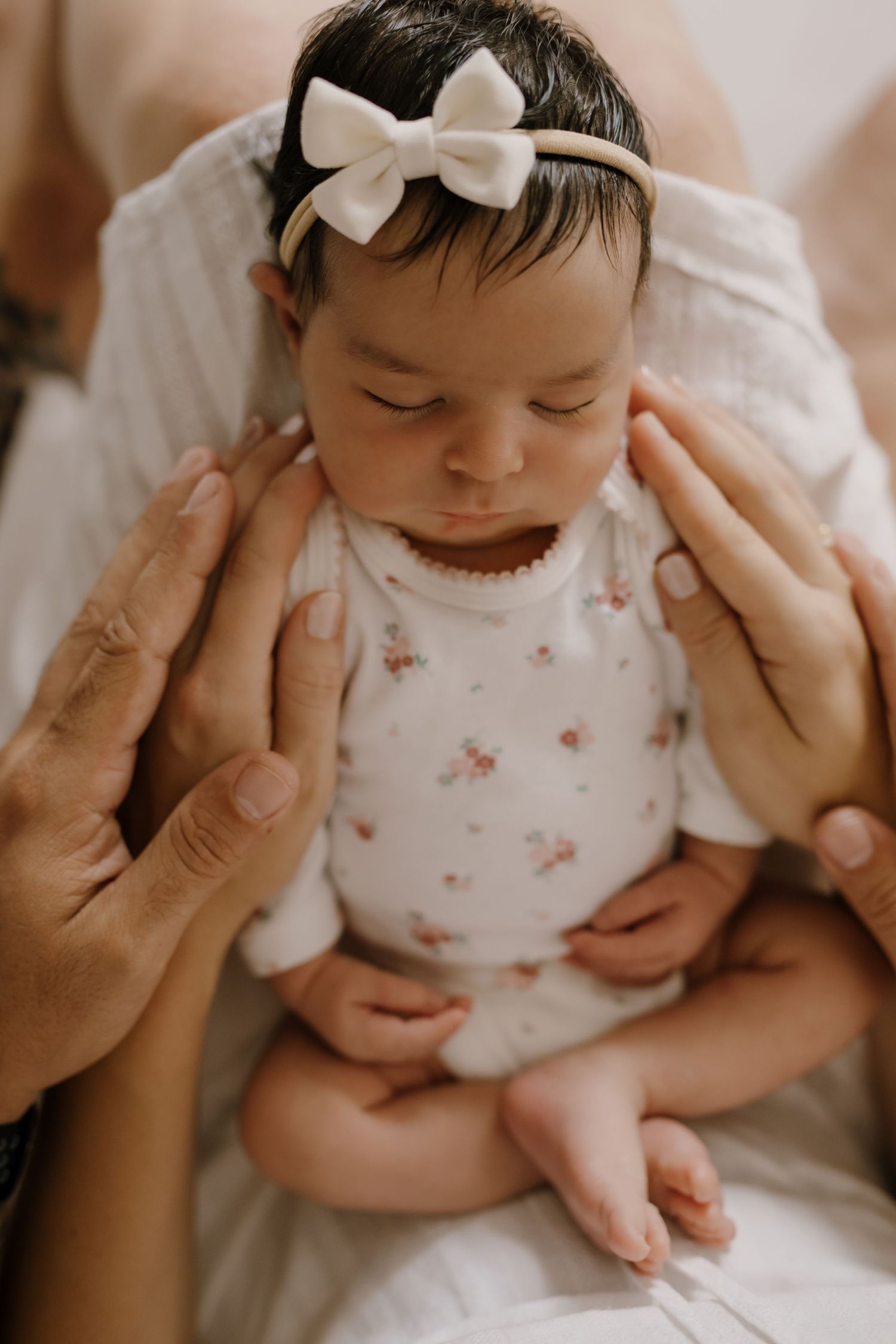 Baby wearing white floral outfit and bow headband cradled in gentle hands.