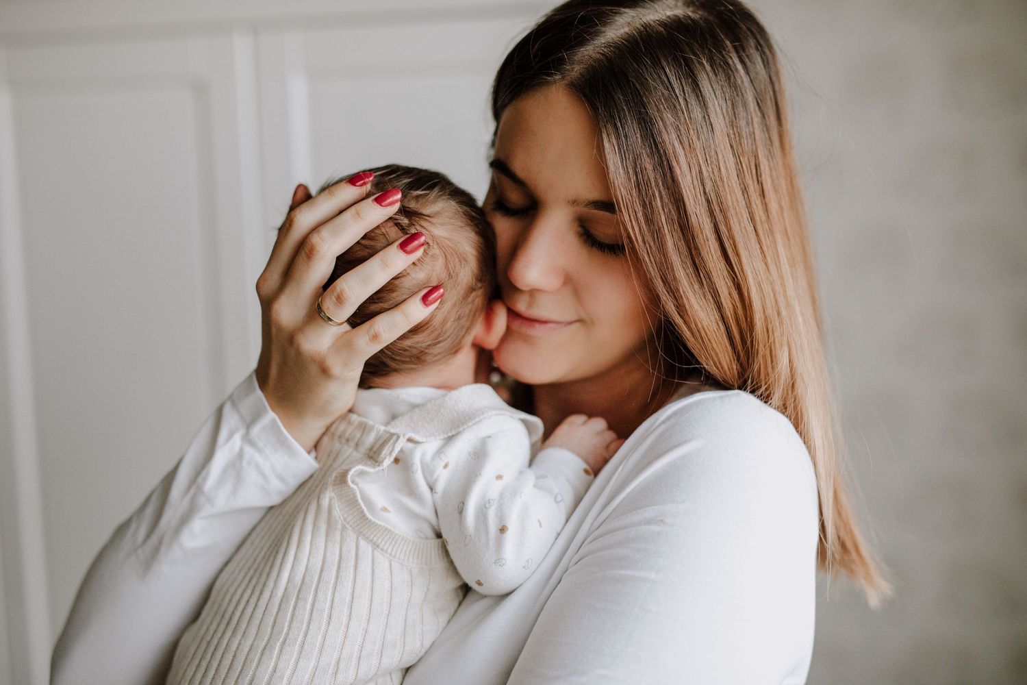 Tender moment between parent and baby in white knit clothing against neutral background.