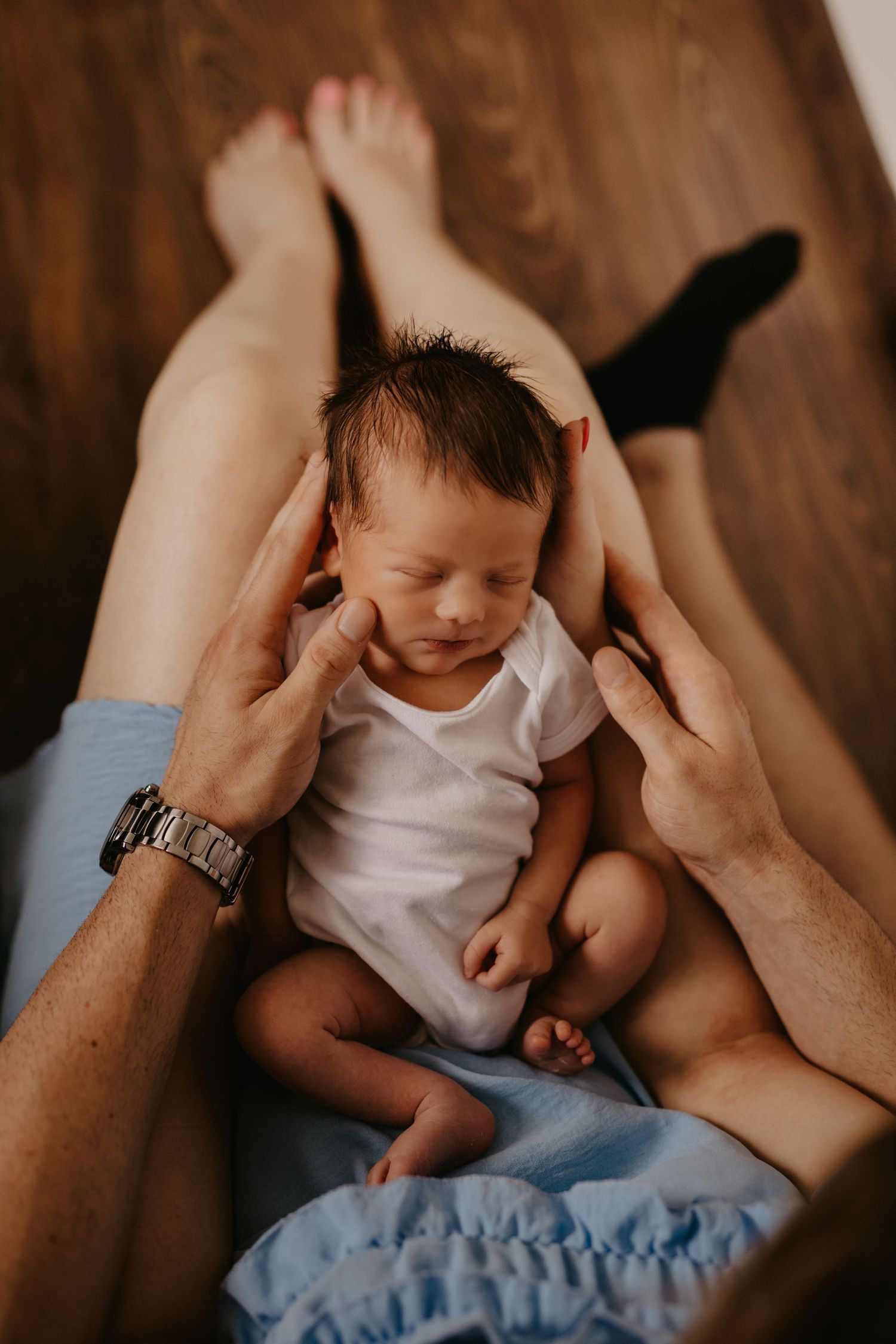 Peaceful newborn in white onesie resting between parents' hands on blue fabric.