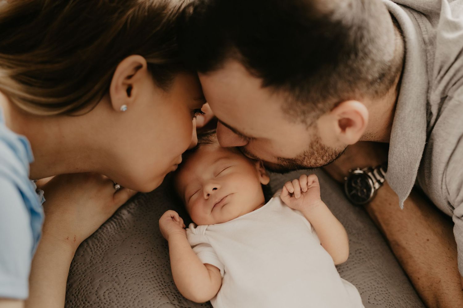 Intimate family moment as parents lovingly embrace their sleeping newborn baby on soft grey bedding.