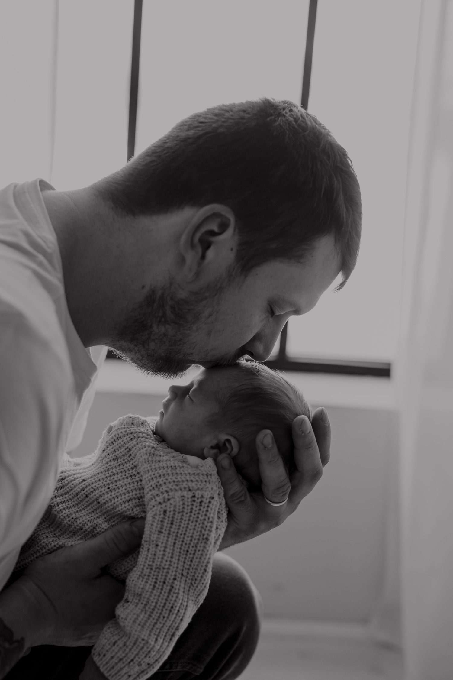 A tender black and white portrait of a father kissing his newborn baby's head.